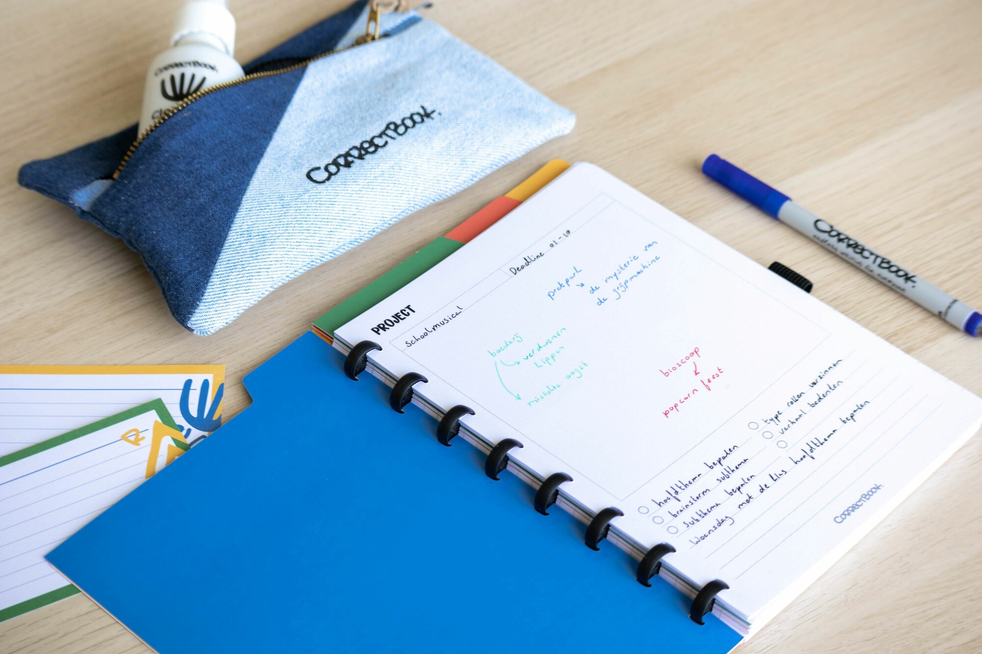 Een open blauw notitieboek van Correctbook ligt op een houten bureau, met een projectplanning ingevuld in gekleurde pennen. Ernaast ligt een denim etui met een flesje Correctbook-spray en een blauwe Correctbook pen.